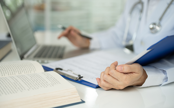 Médico com estetoscópio a consultar um livro e um portátil na secretária